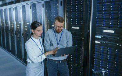 Bearded IT Technician in Glasses with a Laptop Computer and Beautiful Young Engineer Colleague are Talking in Data Center while Working Next to Server Racks. Running Diagnostics or Doing Maintenance Work.